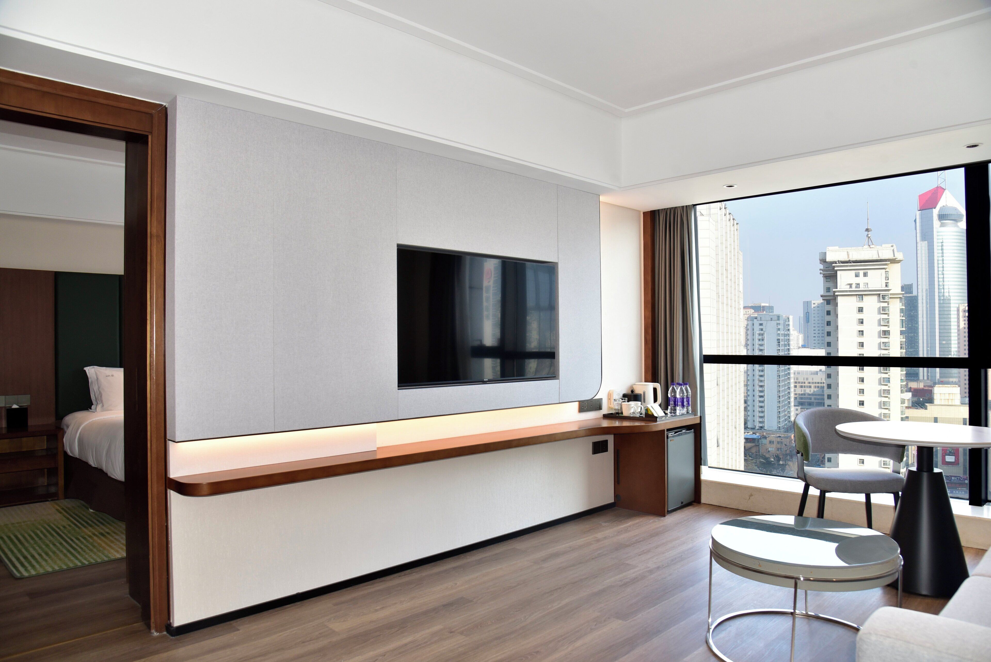 Habitación de hotel moderna con un ventanal grande, un televisor de pantalla plana montado en la pared, una mesita con una silla y una cama con un muro decorado en color verde.