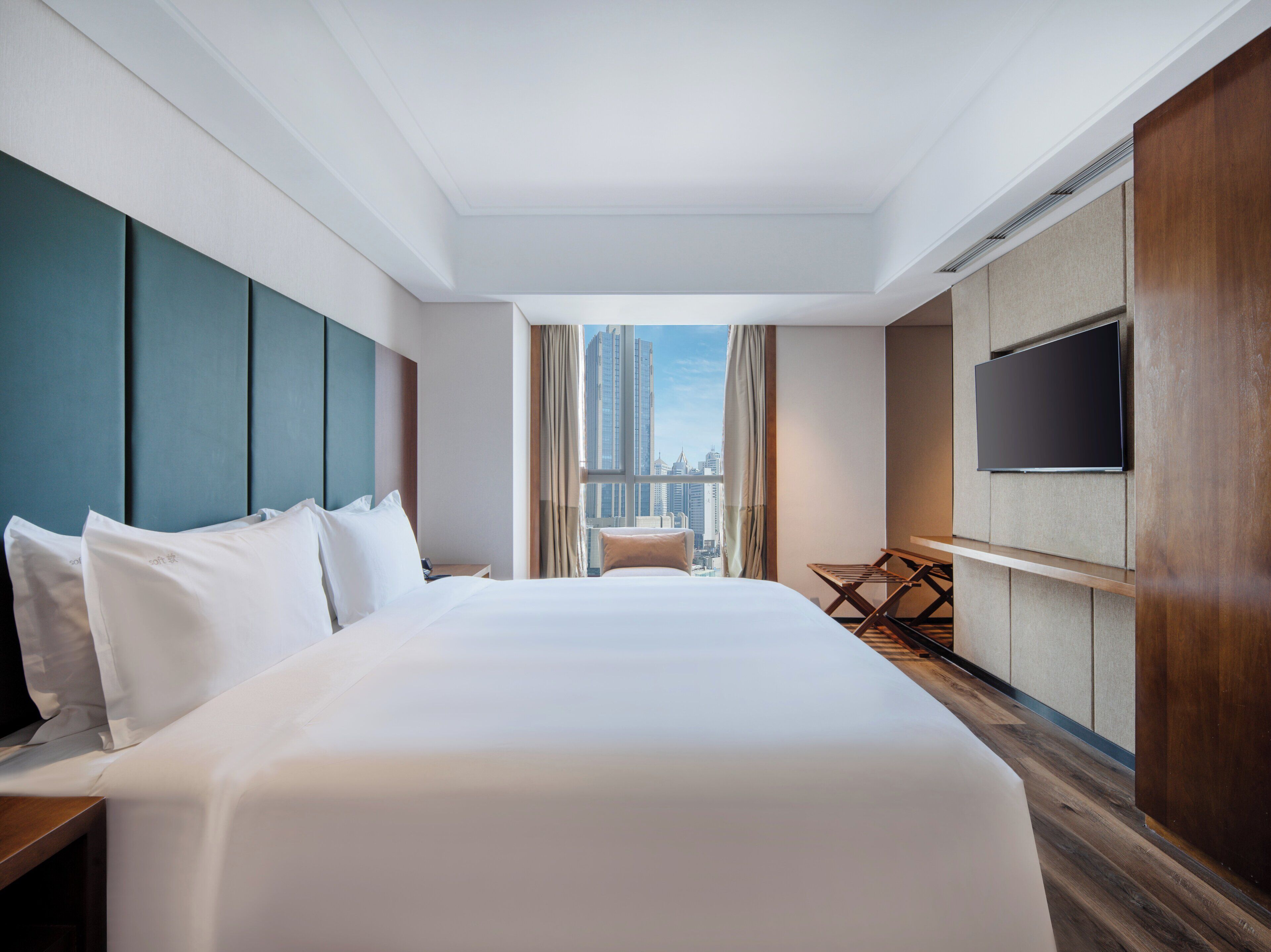 A hotel room with a large bed, a TV mounted on the wall, and a view of the cityscape through the window.