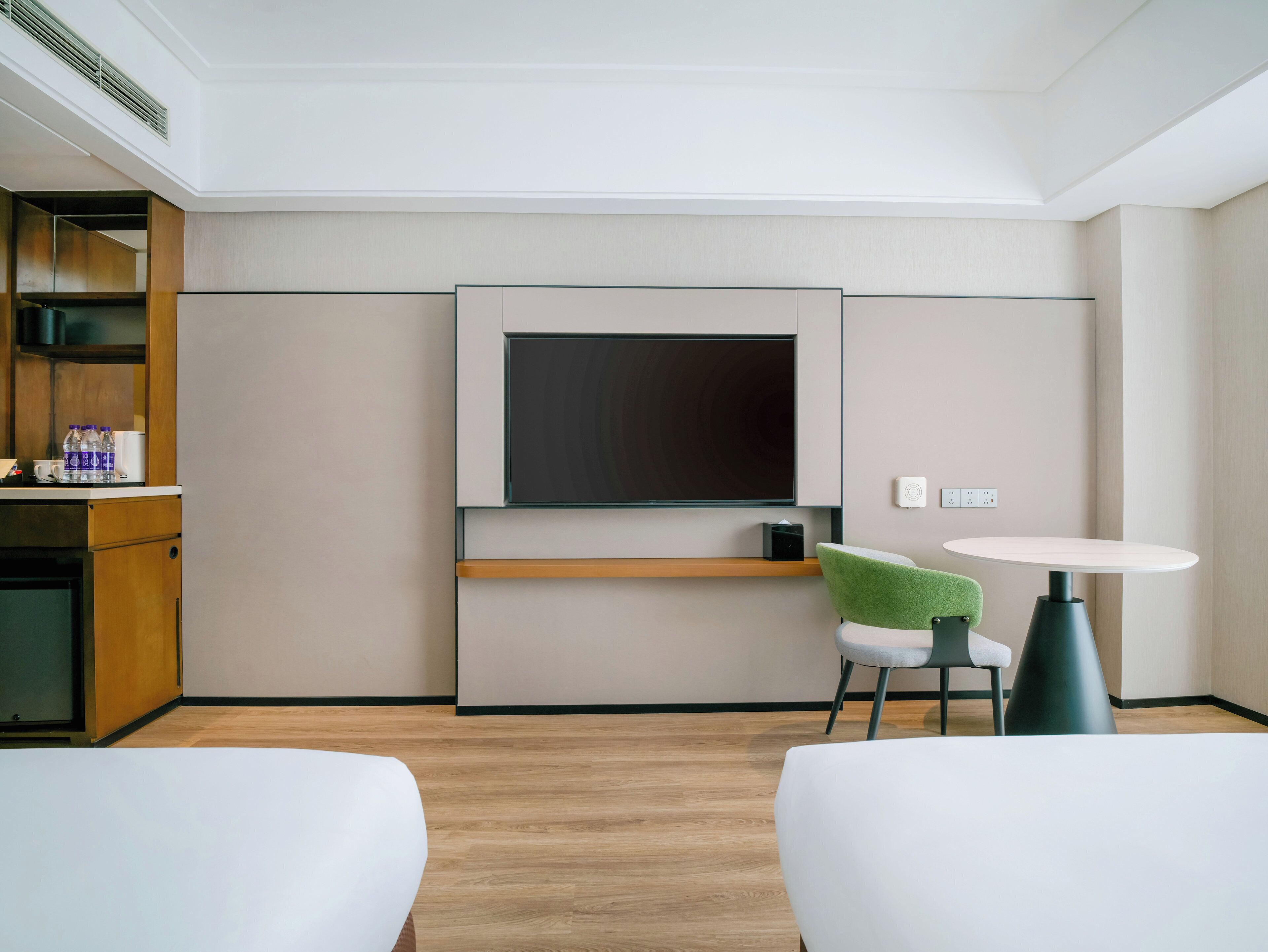 A hotel room with a flat-screen TV, a small table, two chairs, a wooden cabinet, and a shelf with bottled water and a coffee maker.