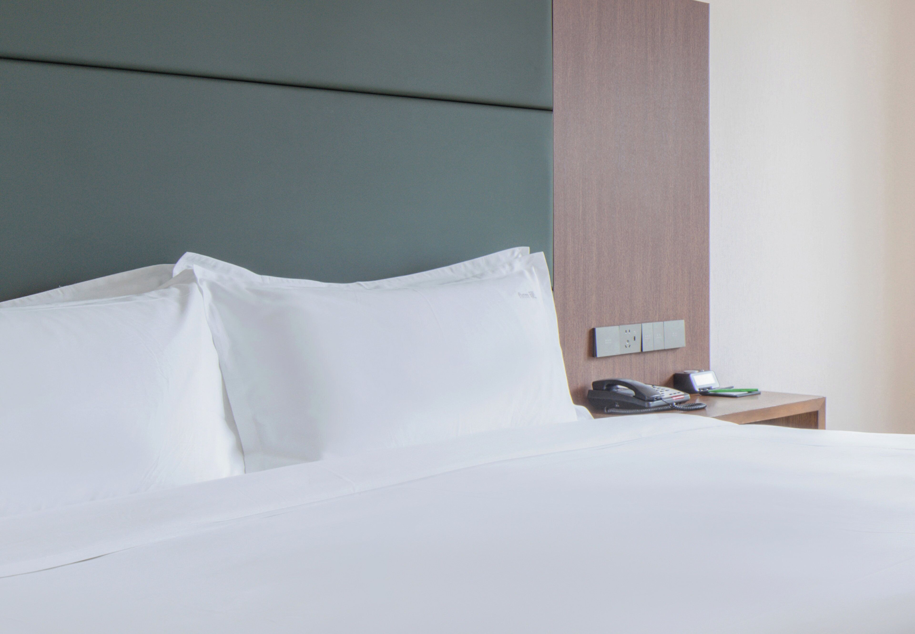 Una cama bien hecha con sábanas blancas, una mesita de noche de madera con un teléfono y un objeto verde, y un interruptor de pared.