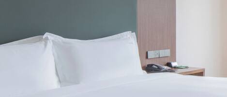 Una cama bien hecha con sábanas blancas, una mesita de noche de madera con un teléfono y un objeto verde, y un interruptor de pared.