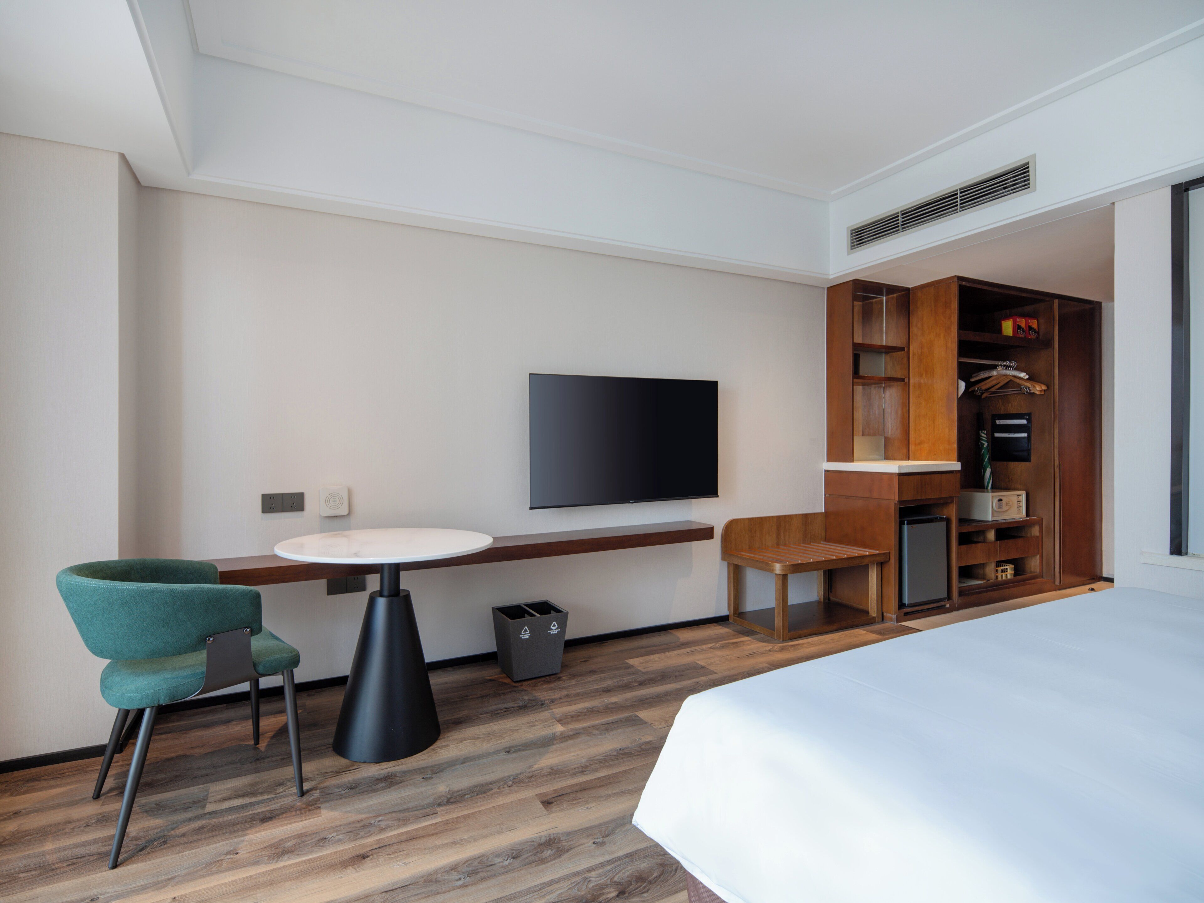 A hotel room with a bed, a TV mounted on the wall, a small table, a chair, and a wooden wardrobe.