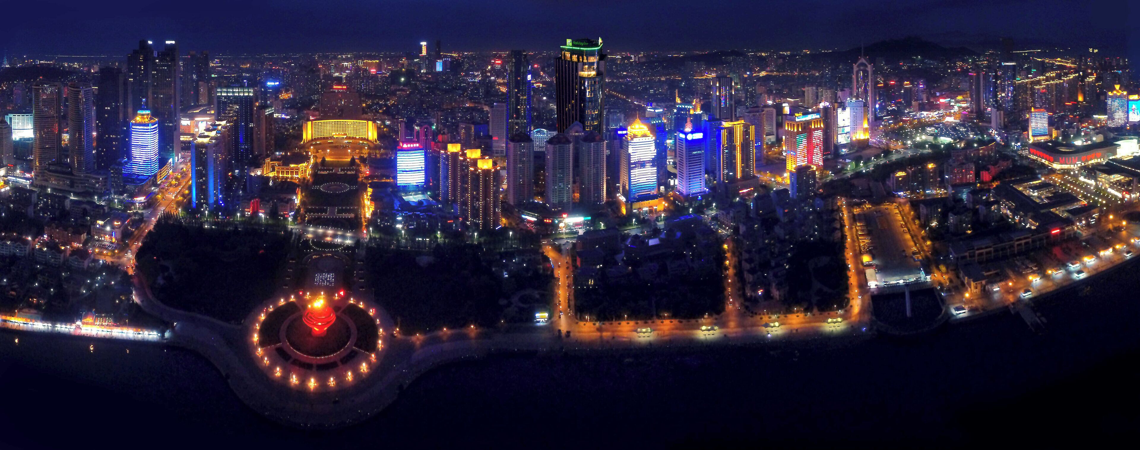 A nighttime cityscape with illuminated buildings and a circular plaza with lights.
