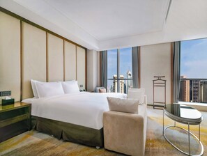 A hotel room with a large bed, a chair, a small table, and a view of the cityscape through large windows. - Holiday Inn Qingdao City Centre by IHG (Qingdao)