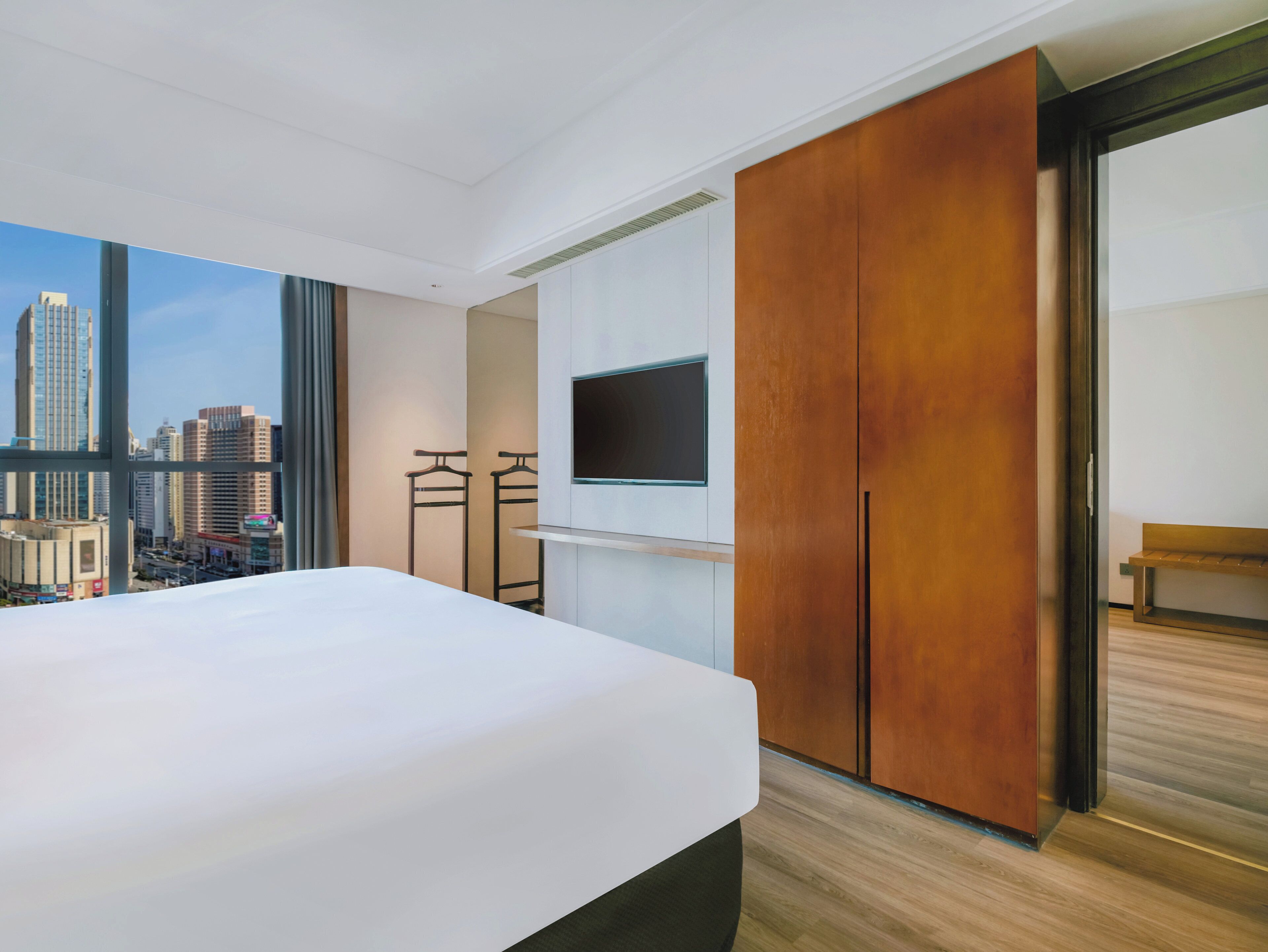 A modern hotel room with a large bed, a TV mounted on the wall, and a view of a cityscape through large windows.