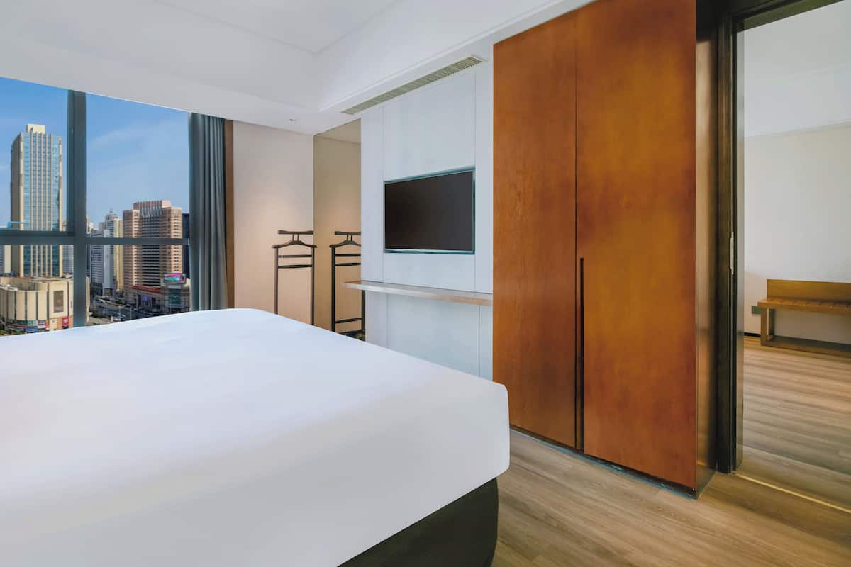 A modern hotel room with a large bed, a TV mounted on the wall, and a view of a cityscape through large windows.