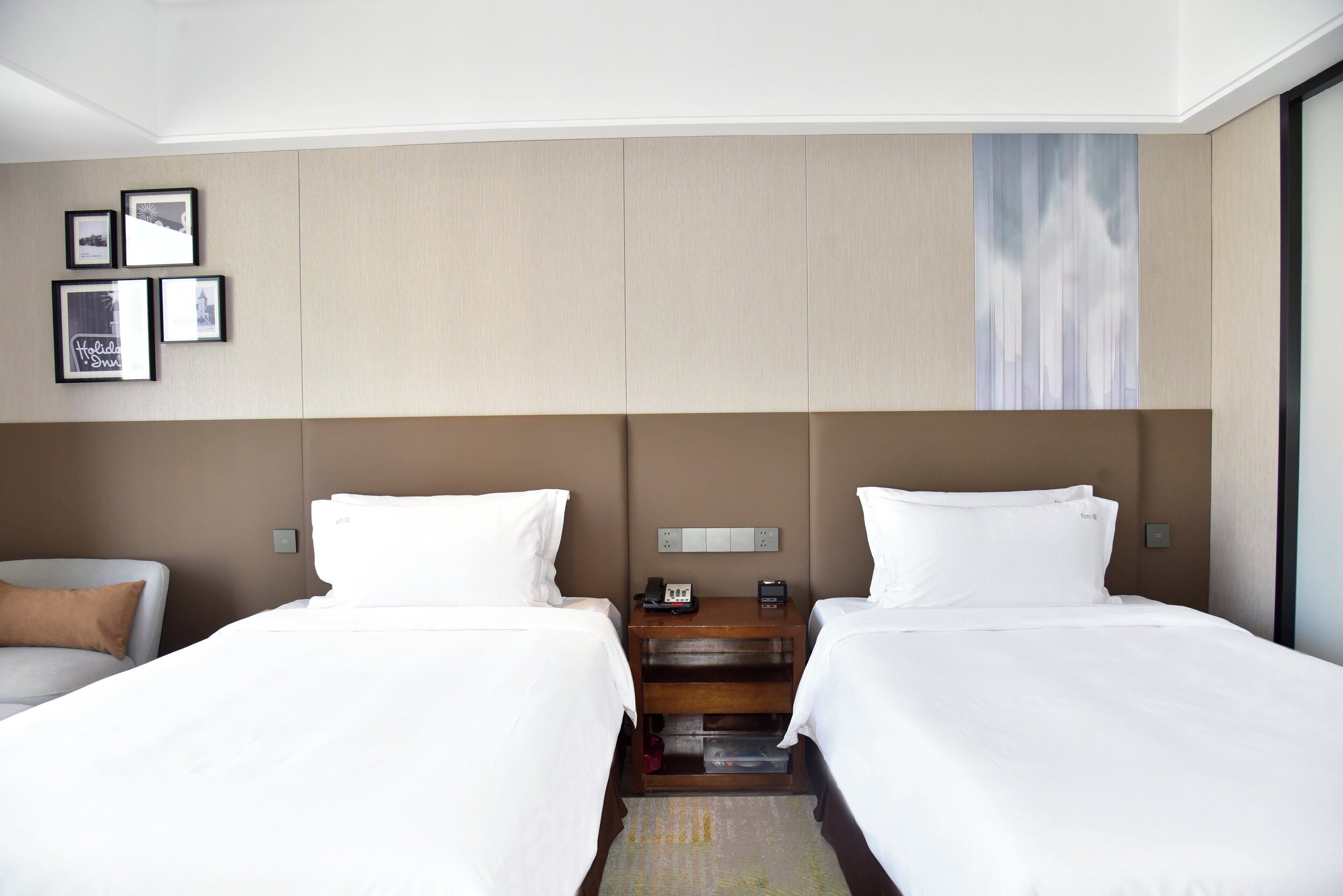 A hotel room with two beds, a nightstand, a lamp, and a wall-mounted artwork.