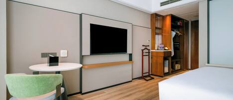 Ein Hotelzimmer mit einem Bett, einem an der Wand befestigten Fernseher, einem kleinen runden Tisch, einem grünen Stuhl, einem Kleiderschrank und Holzboden.