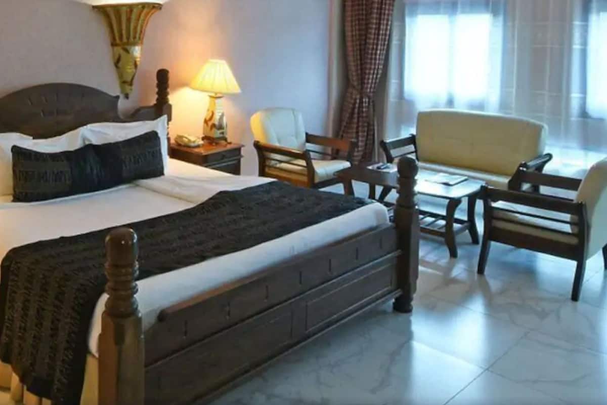 Habitación de hotel con una cama grande, dos mesitas de noche con lámparas, una silla, una pequeña mesa y una ventana con cortinas.