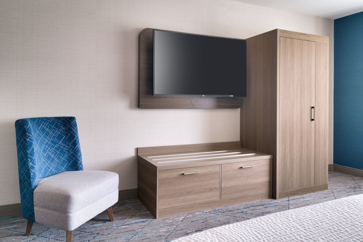 A hotel room with a flat-screen TV mounted on the wall, a wooden wardrobe with two drawers, a blue patterned armchair, and a patterned carpet.