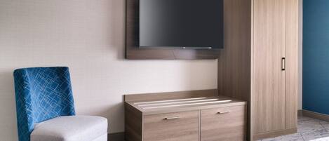 Una habitación de hotel con un televisor de pantalla plana montado en la pared, un armario de madera con dos cajones, un sillón azul estampado y una alfombra con diseño.