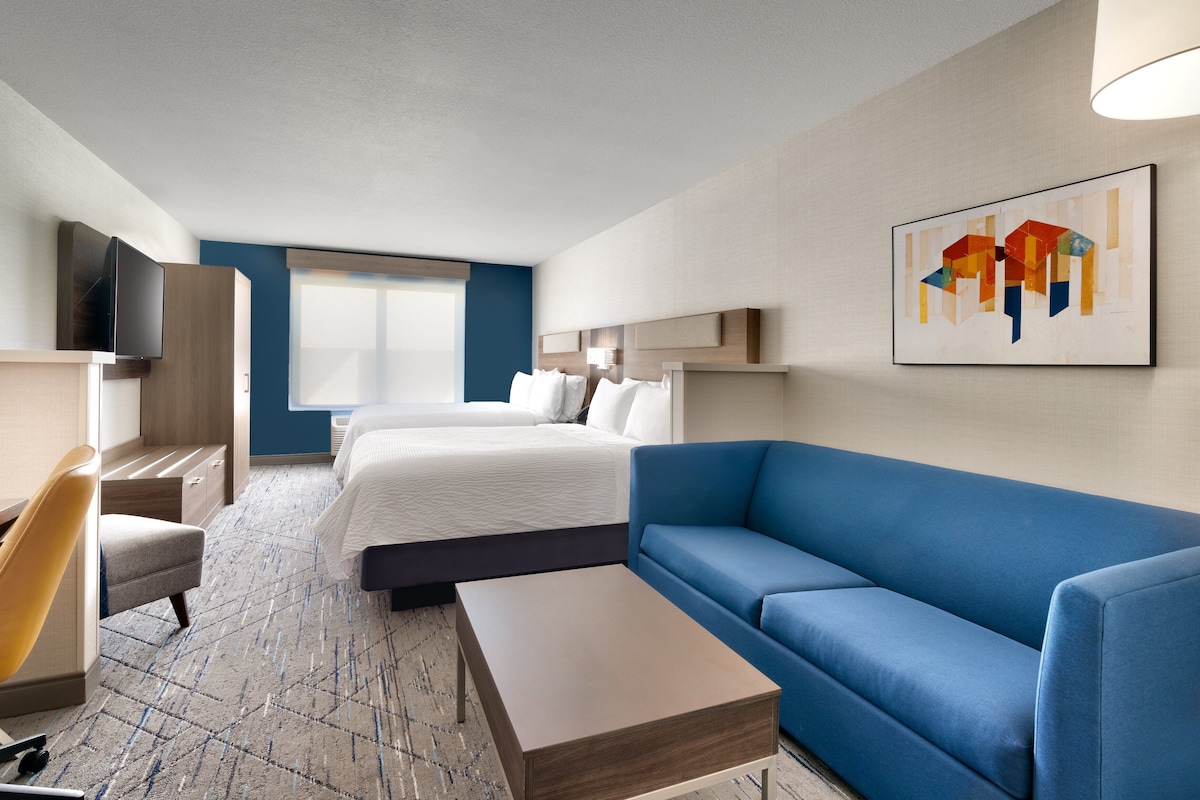 Habitación de hotel con dos camas, un sofá azul, una mesita de centro de madera, un televisor y un cuadro en la pared.