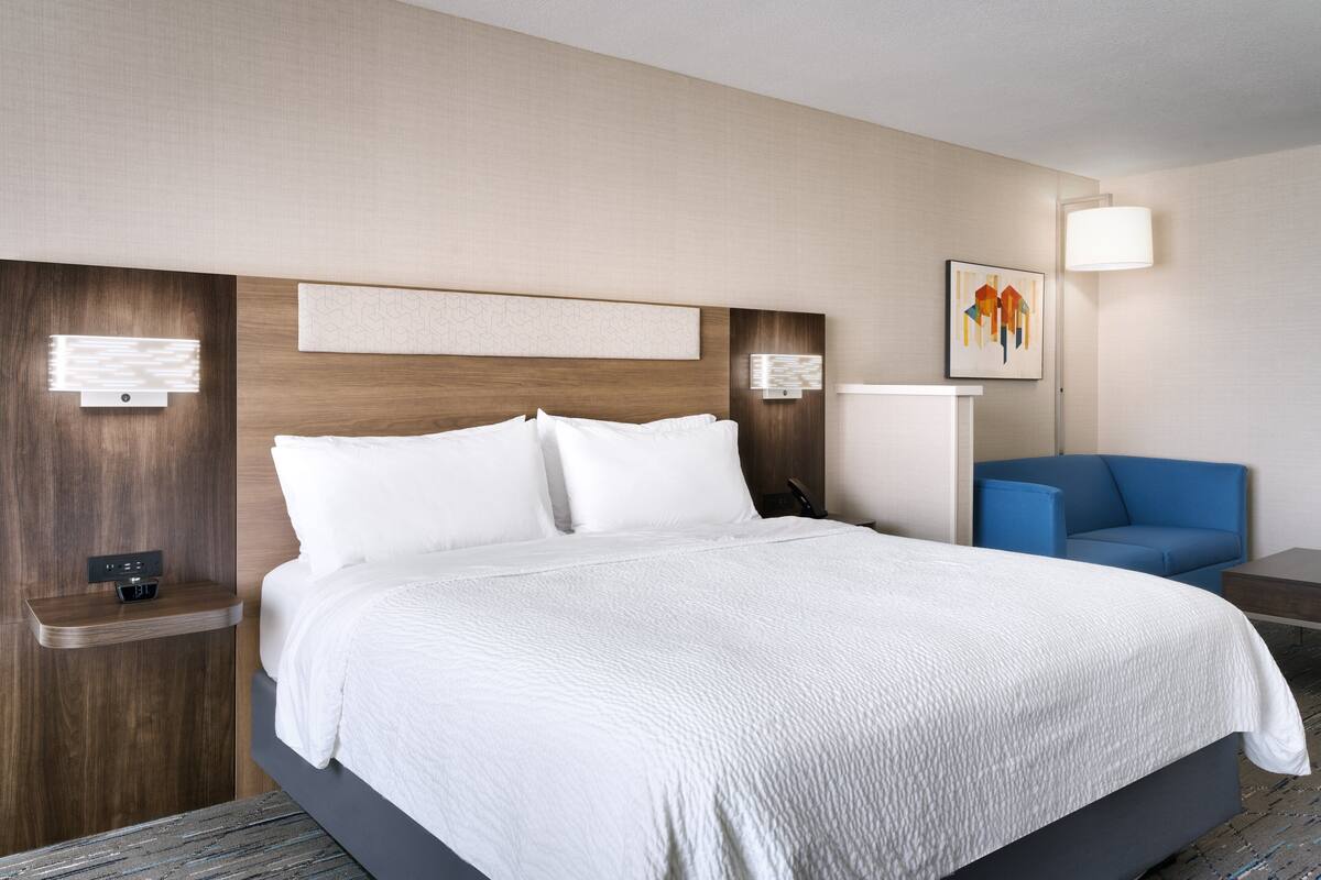 A modern hotel room with a large bed, a bedside table with a lamp, a blue armchair, and a wall-mounted artwork.