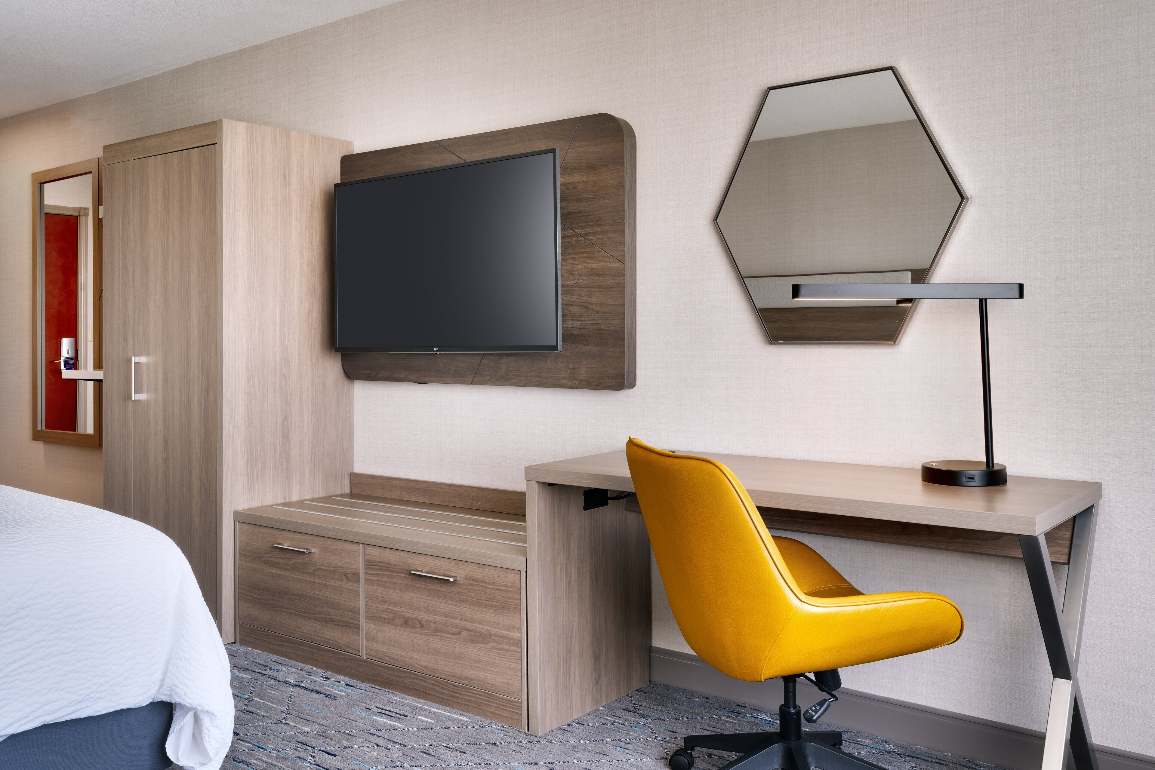 A modern hotel room with a bed, a desk with a chair, a flat-screen TV, and a large mirror.