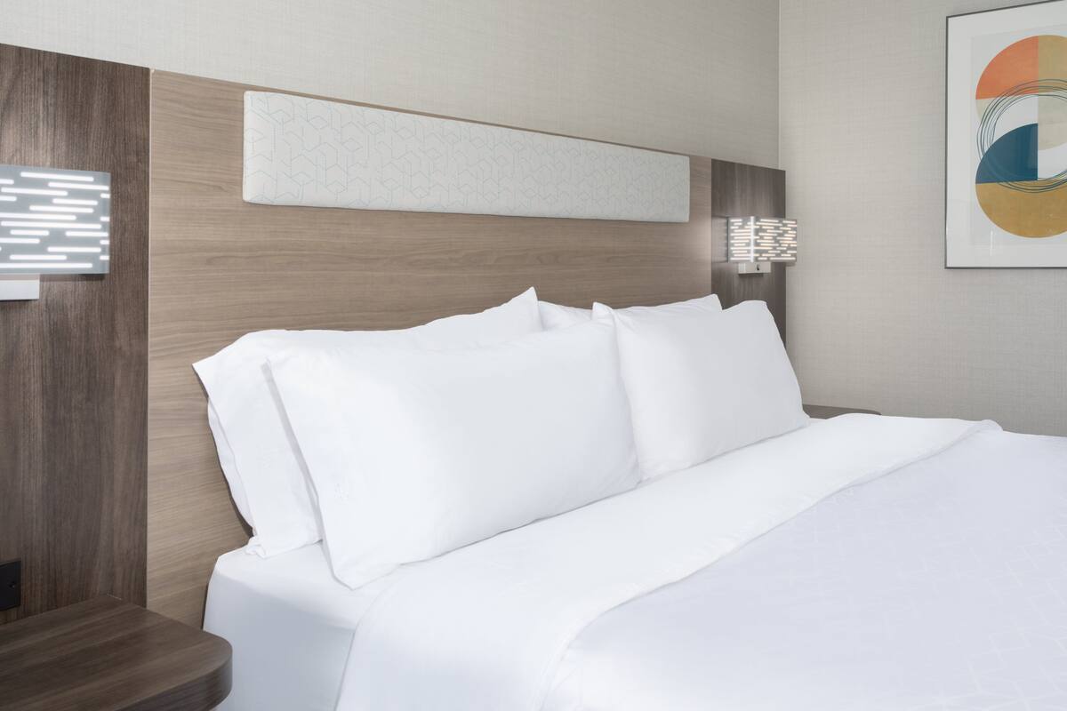 A modern hotel room with a neatly made bed, a wooden headboard, a wall-mounted light, and a framed abstract artwork.