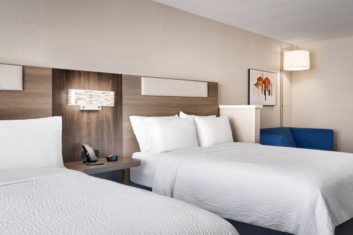 A hotel room with two beds, a bedside table with a lamp, a clock, and artwork on the wall.