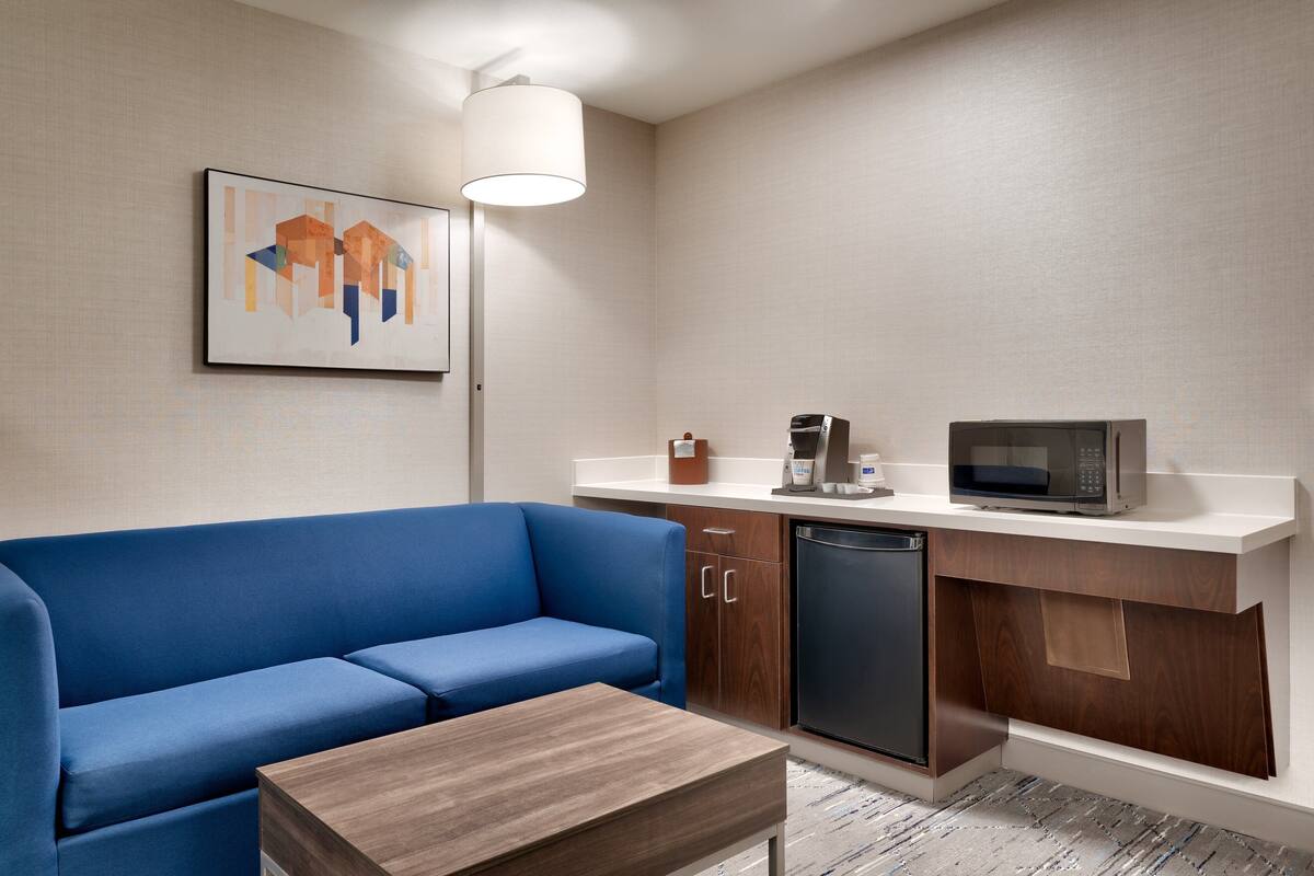 A compact living space with a blue sofa, a wooden coffee table, a kitchen area with a microwave and coffee maker, and a wall-mounted abstract painting.