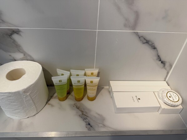 Un baño con piso de mármol, un rollo de papel higiénico y varios artículos de tocador, incluyendo jabón de baño, champú, acondicionador y una caja de productos de higiene personal.