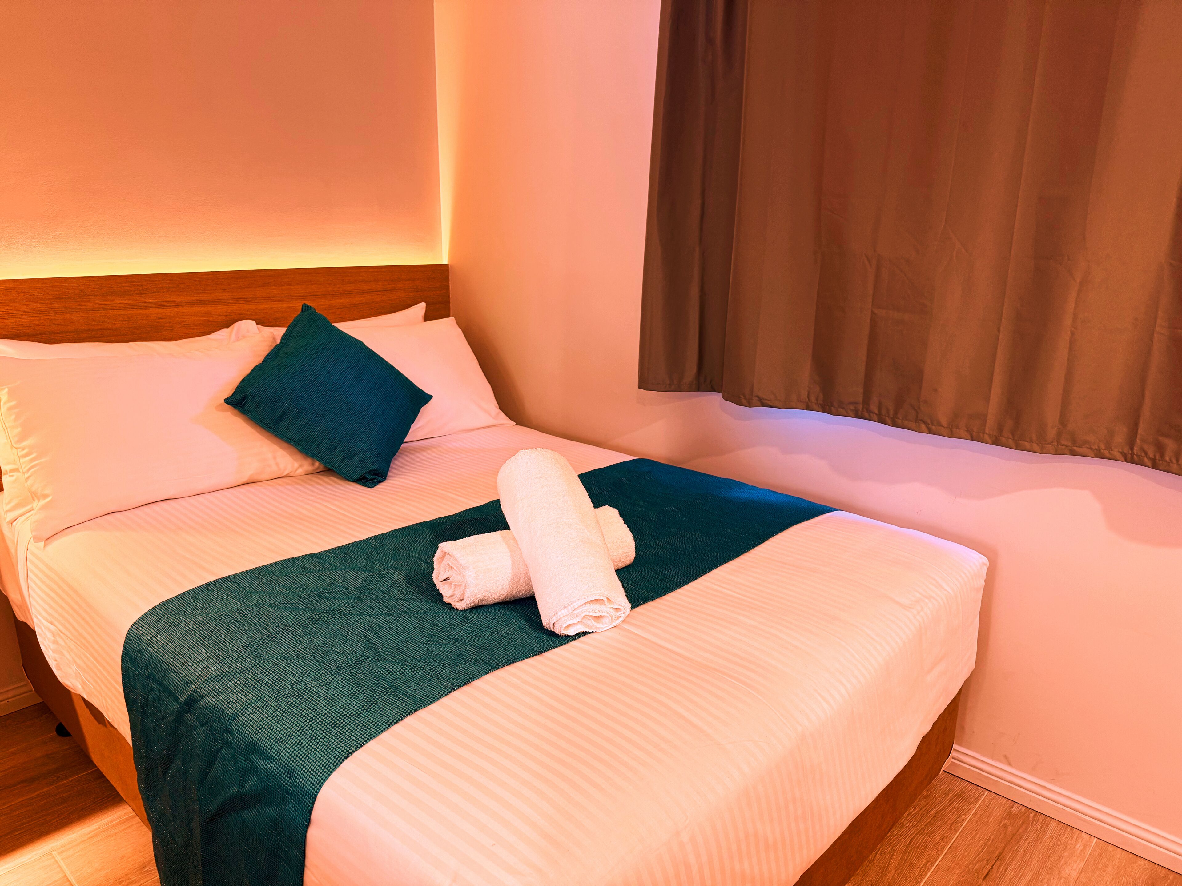 Una cama bien hecha con una manta verde, una toalla enrollada y una almohada oscura.
