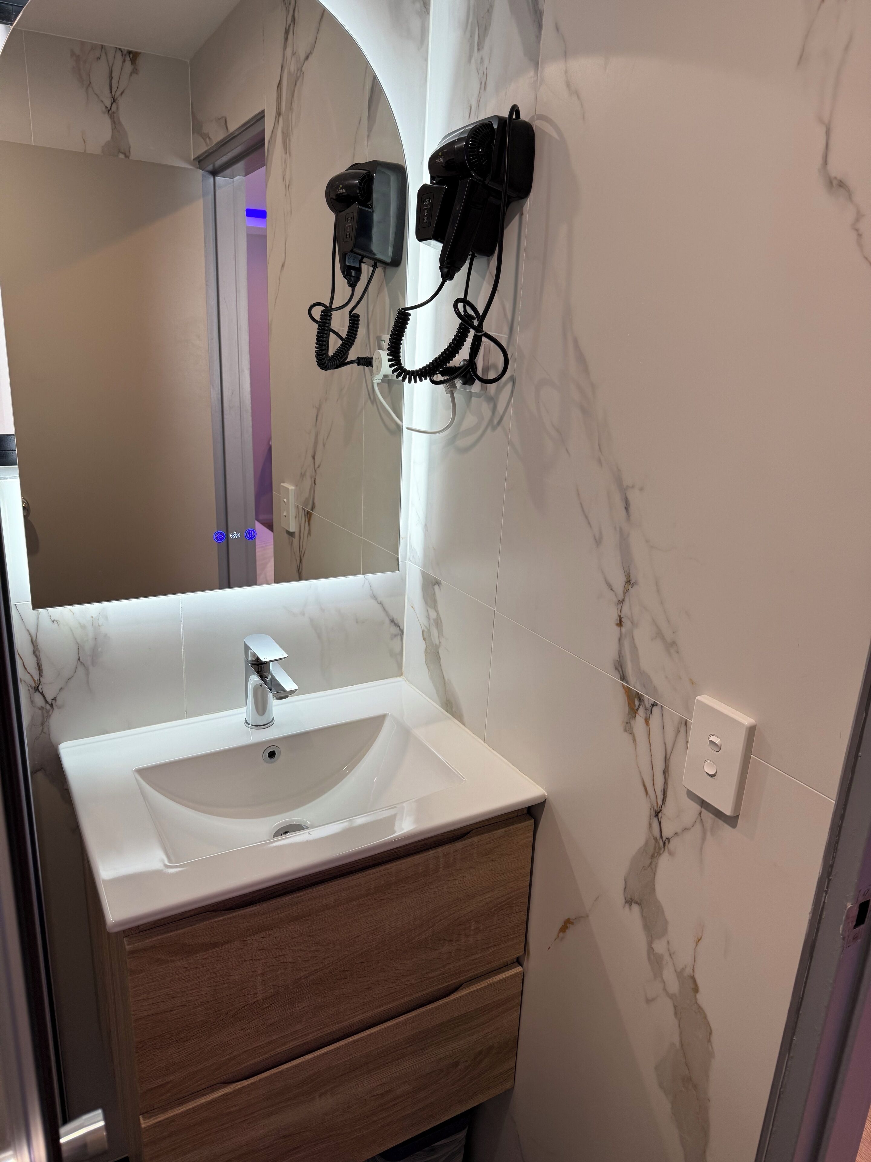Un baño con pared de mármol, un lavamanos blanco, un mueble de tocador de madera y dos secadores de pelo montados en la pared.