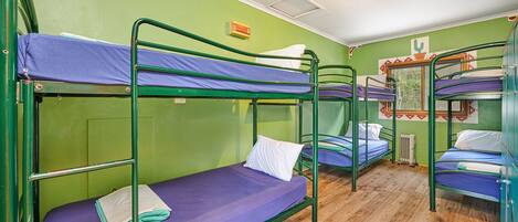 A dormitory room with bunk beds, green walls, wooden flooring, and a small window with a view of trees.