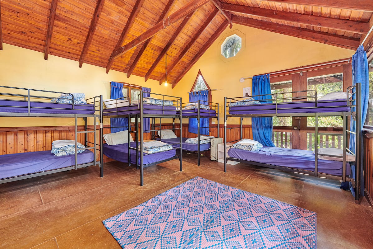 A room with four bunk beds, wooden beams on the ceiling, and a large window with curtains.