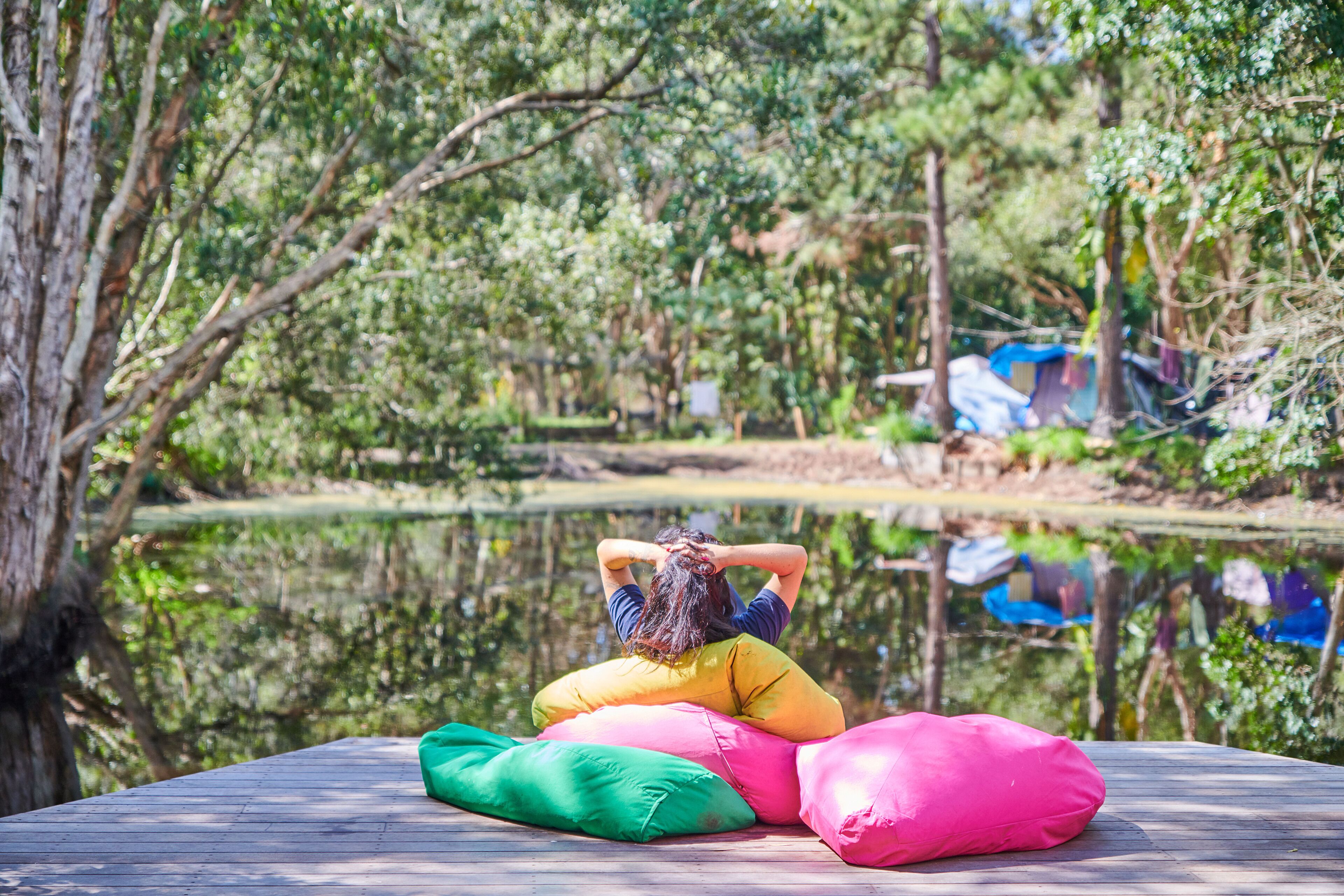 A person sitting on a wooden deck with colorful cushions, surrounded by a natural environment with trees and a body of water.