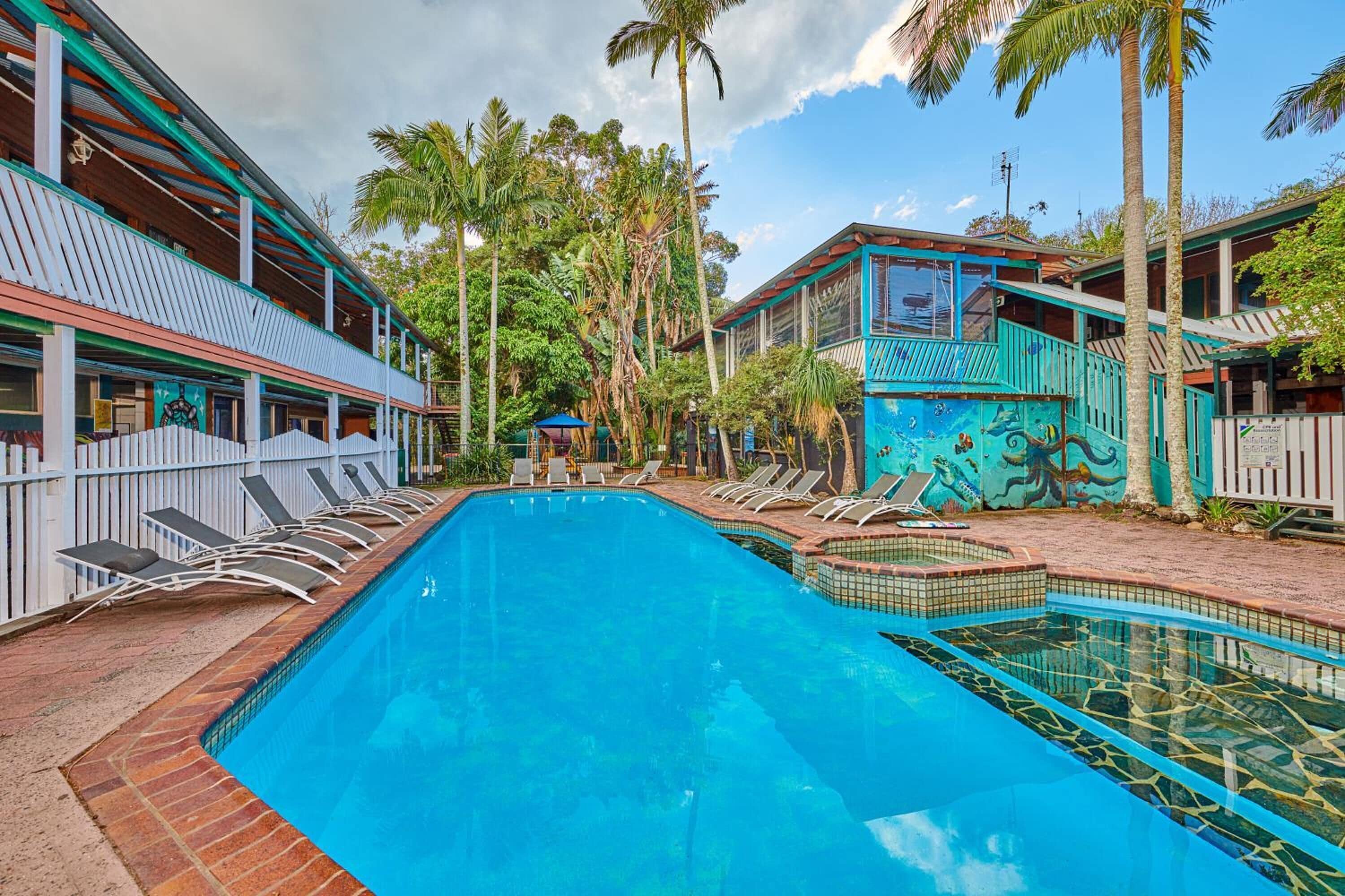 Una piscina con sillones de descanso, rodeada de edificios con temática tropical, incluyendo palmeras y un mural.