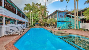 Una piscina con sillones de descanso, rodeada de edificios con temática tropical, incluyendo palmeras y un mural.