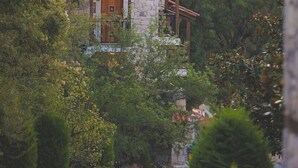 Una casa de piedra con techo de tejas rojas, rodeada de abundante vegetación y montañas al fondo.