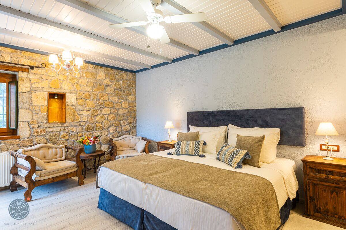 A bedroom with a stone wall, a large bed with multiple pillows, a wooden chair, and a small table with a vase of flowers.