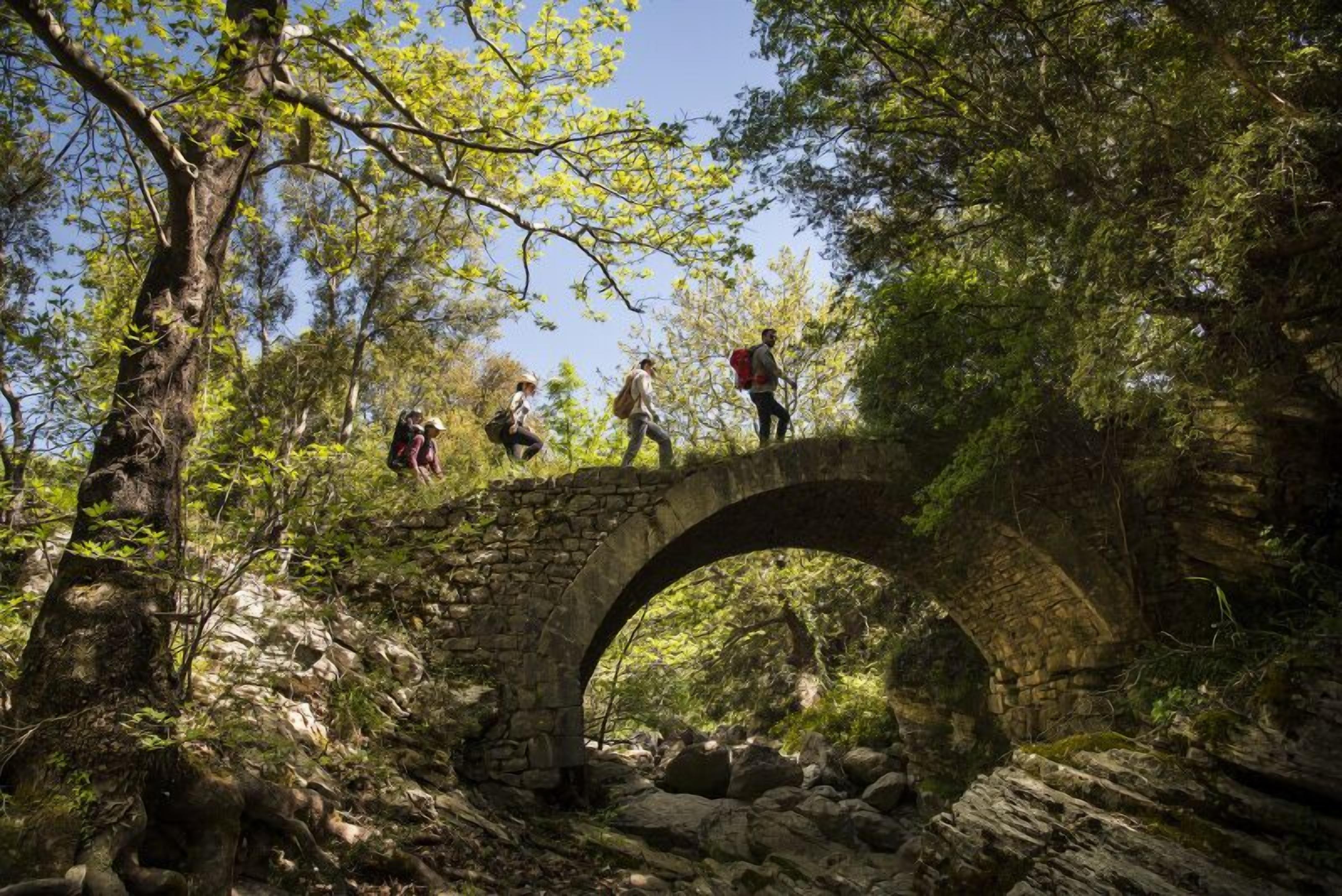 Un puente de piedra en un área boscosa, con excursionistas cruzándolo.