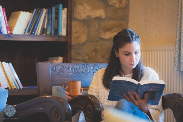 A person sitting on a chair reading a book in a room with a bookshelf and a stone wall. - Abeliona Retreat (Oichalia)