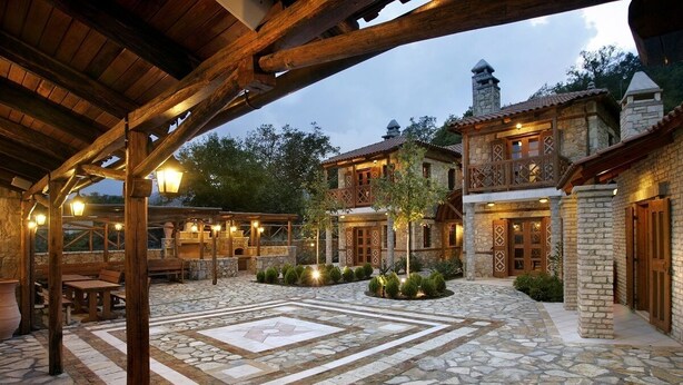 Un patio de piedra con mobiliario de madera y suelo de mosaico, rodeado de edificios de piedra con balcones e iluminación cálida.