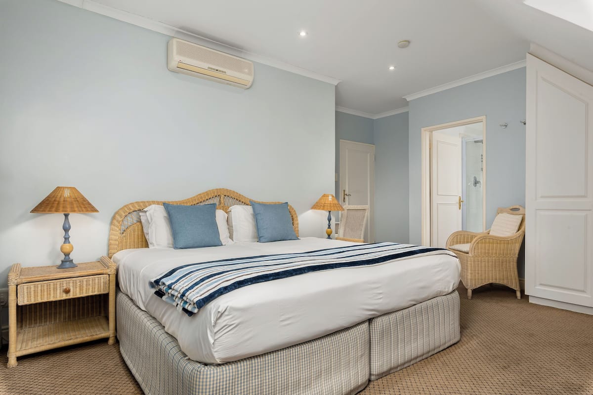 A bedroom with a large bed, two nightstands, a wicker chair, and a door leading to another room.