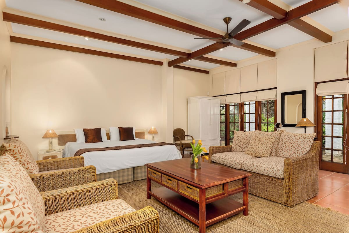 A hotel room with a bed, a sofa, a coffee table, wicker furniture, a ceiling fan, and large windows.