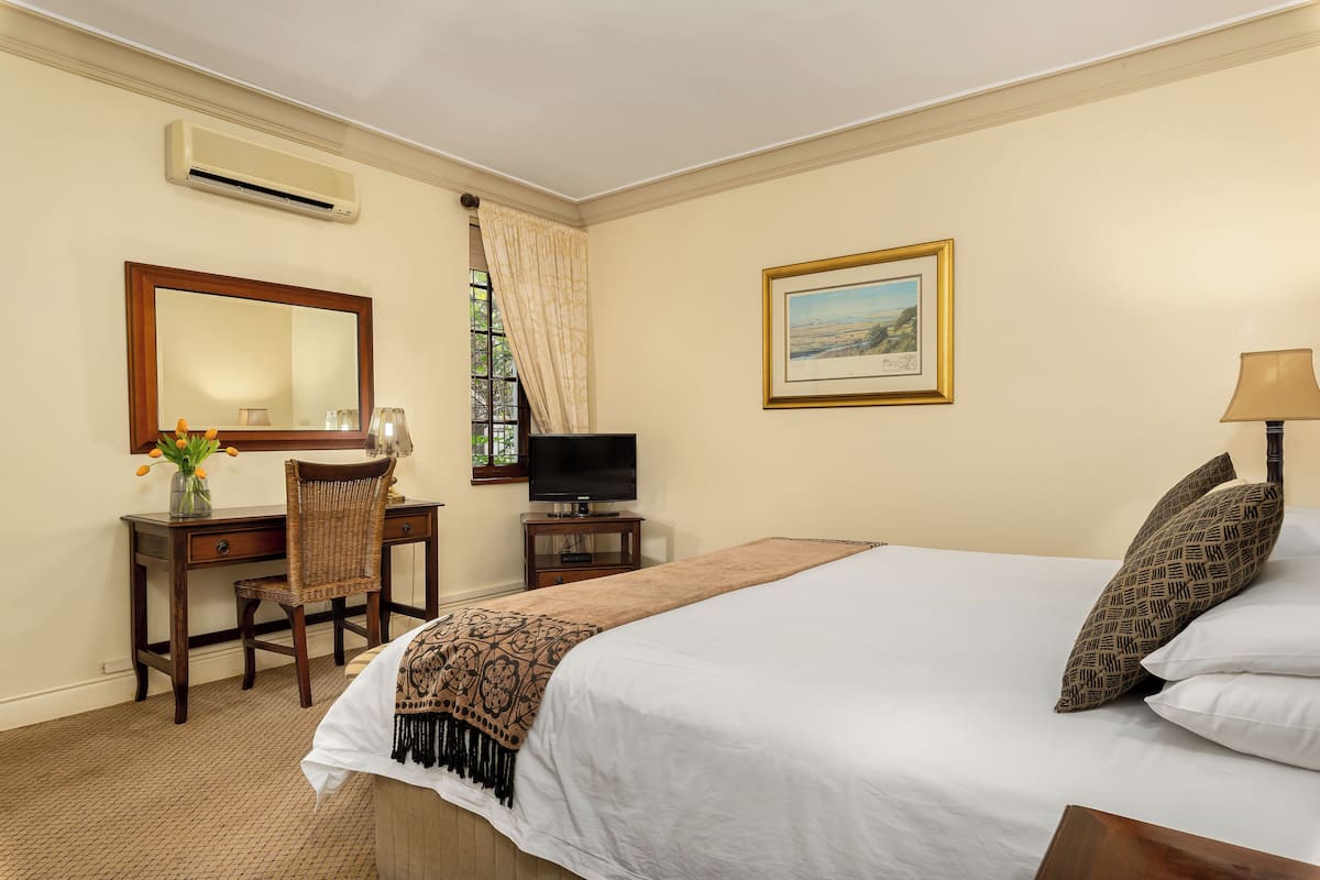 A hotel room with a bed, a desk with a chair, a television, a framed picture on the wall, and an air conditioning unit.
