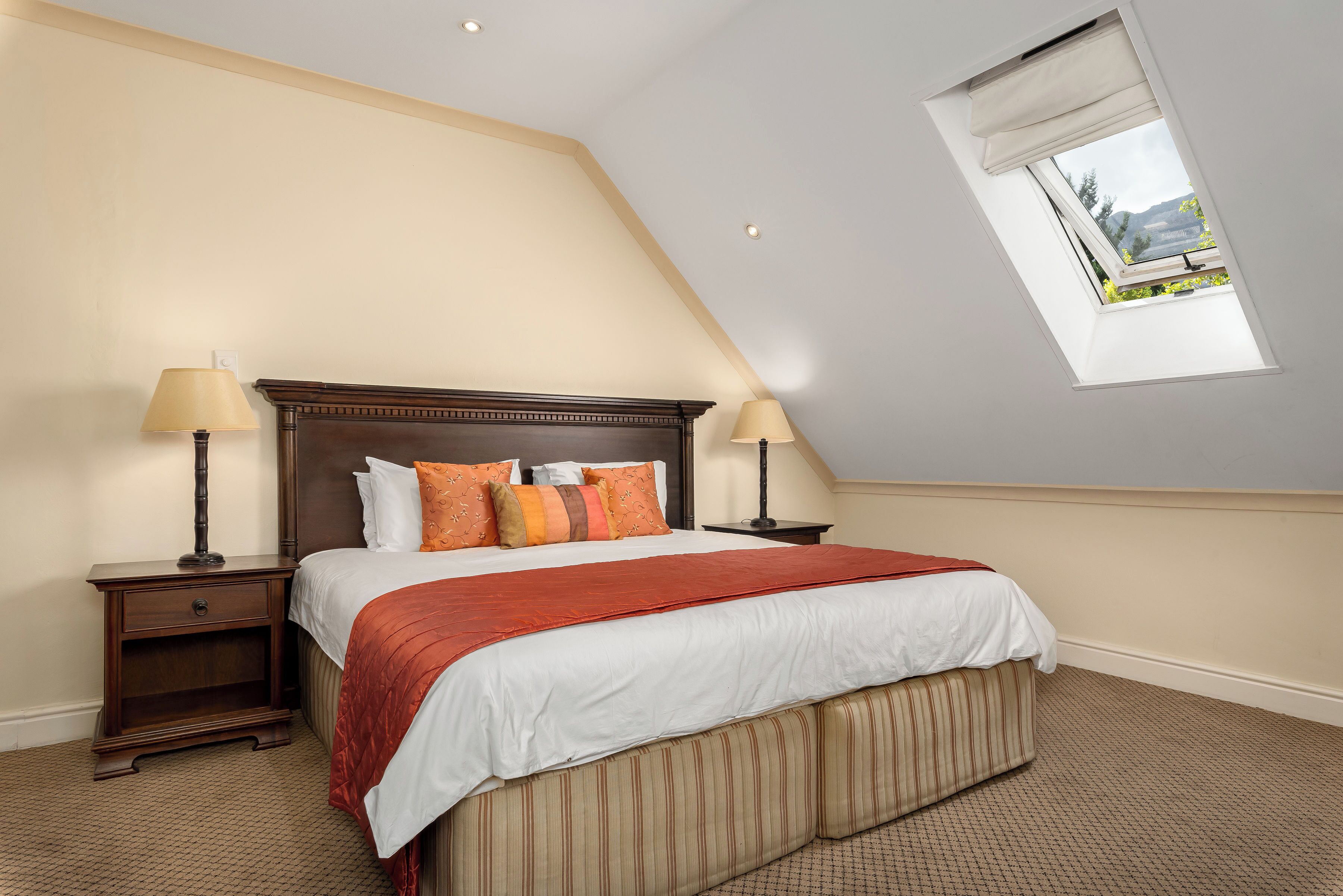 A bedroom with a sloped ceiling, a large bed with a red blanket, two bedside tables with lamps, and a window with a view of greenery.