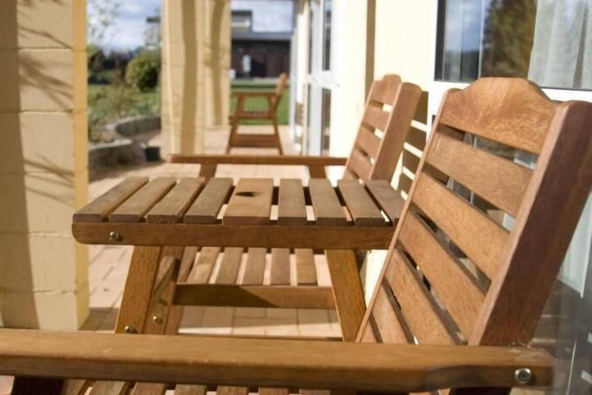 A wooden bench with a backrest and armrests, positioned on a patio with a view of a building and greenery in the background.
