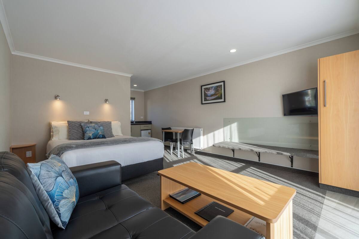 A modern hotel room with a bed, a sofa, a wooden coffee table, a desk, a chair, a TV, and a wardrobe.