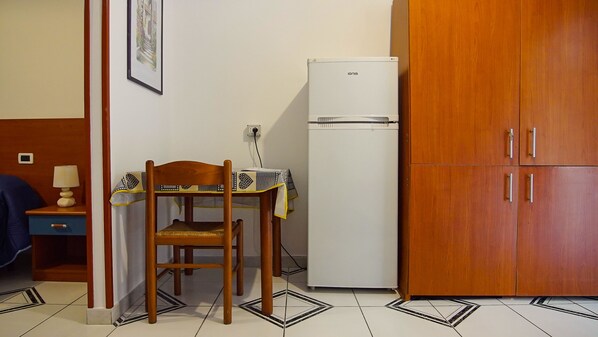 A small kitchenette with a wooden chair, a table with a cloth, a white refrigerator, and wooden cabinets. - Baia di Naxos (Giardini Naxos)