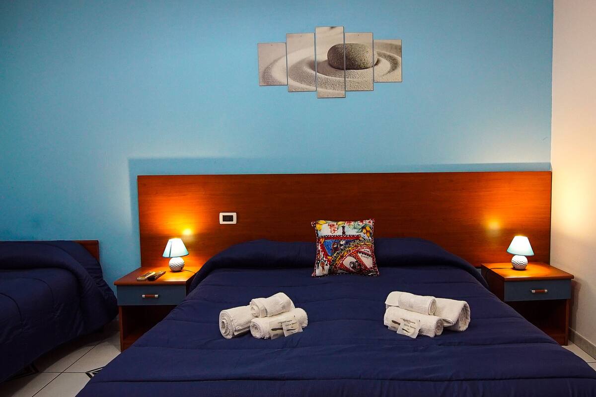 Ett hotellrum med en blå vägg, en sänggavel i trä, två sängbord med lampor, en säng med blått sänglinne, två hoprullade handdukar och en färgglad kudde.