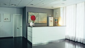 Een receptiegedeelte met een wit bureau, een groot abstract schilderij, een prikbord en een raam met dunne gordijnen.