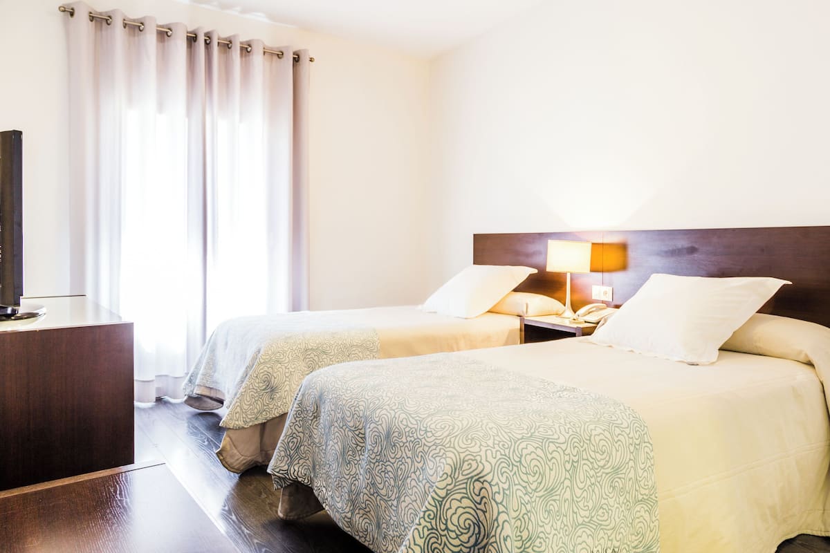 A hotel room with two beds, a TV, a nightstand with a lamp, and a window with curtains.