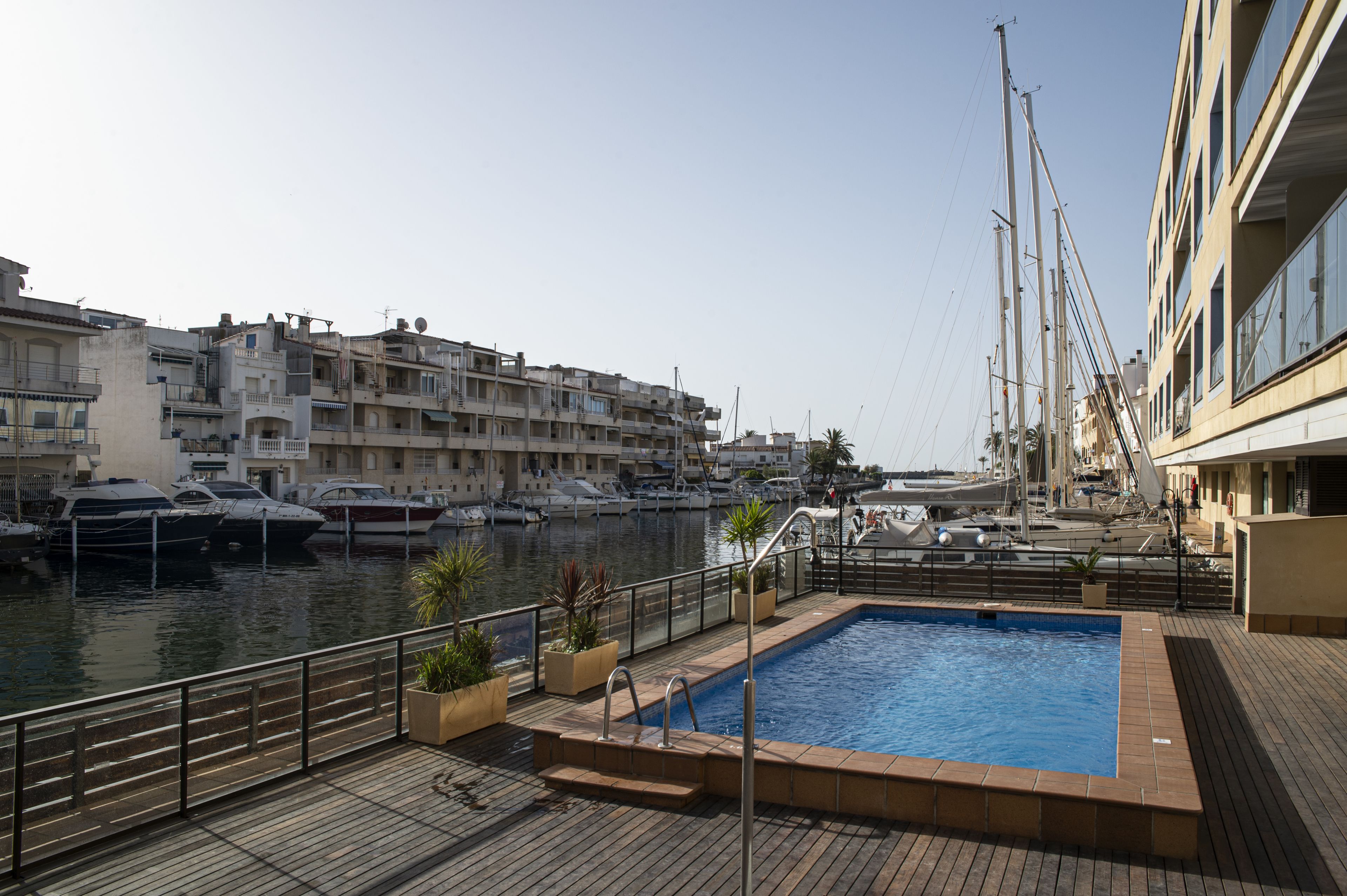 A marina with boats docked, a swimming pool, and a building with balconies overlooking the water.