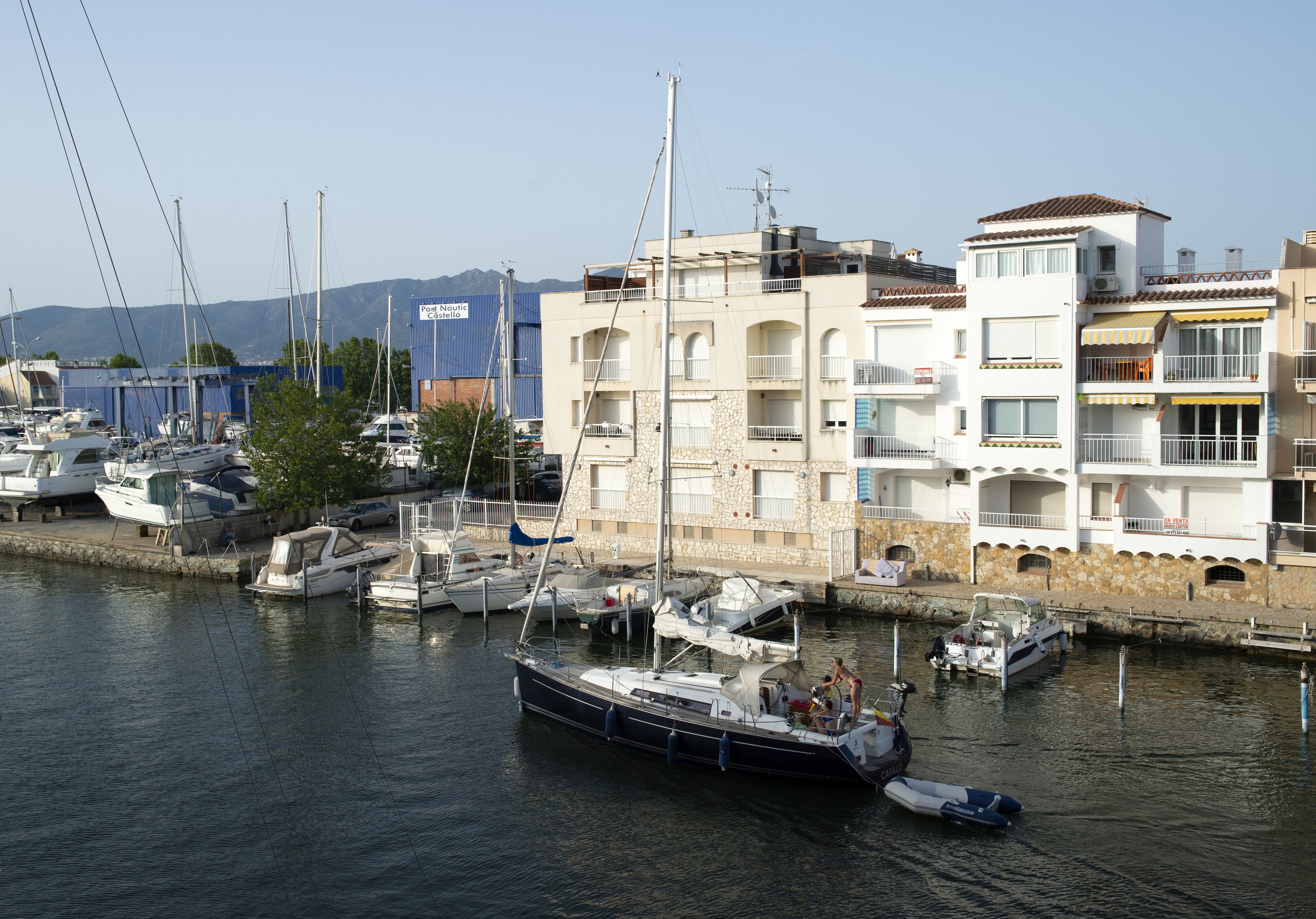 A marina with boats docked, a building with multiple balconies, and a clear sky.