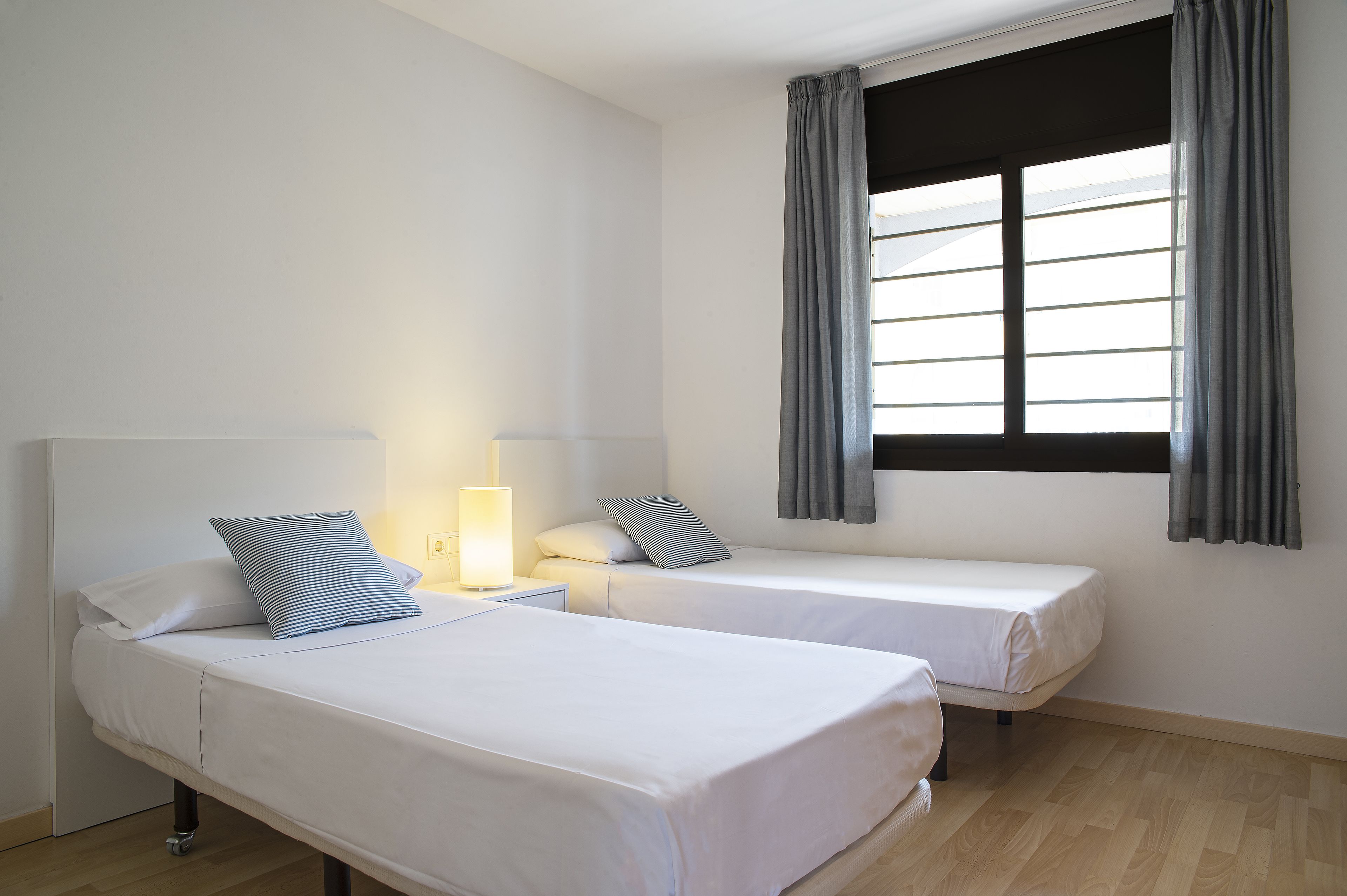 A room with two single beds, a nightstand with a lamp, and a window with curtains.