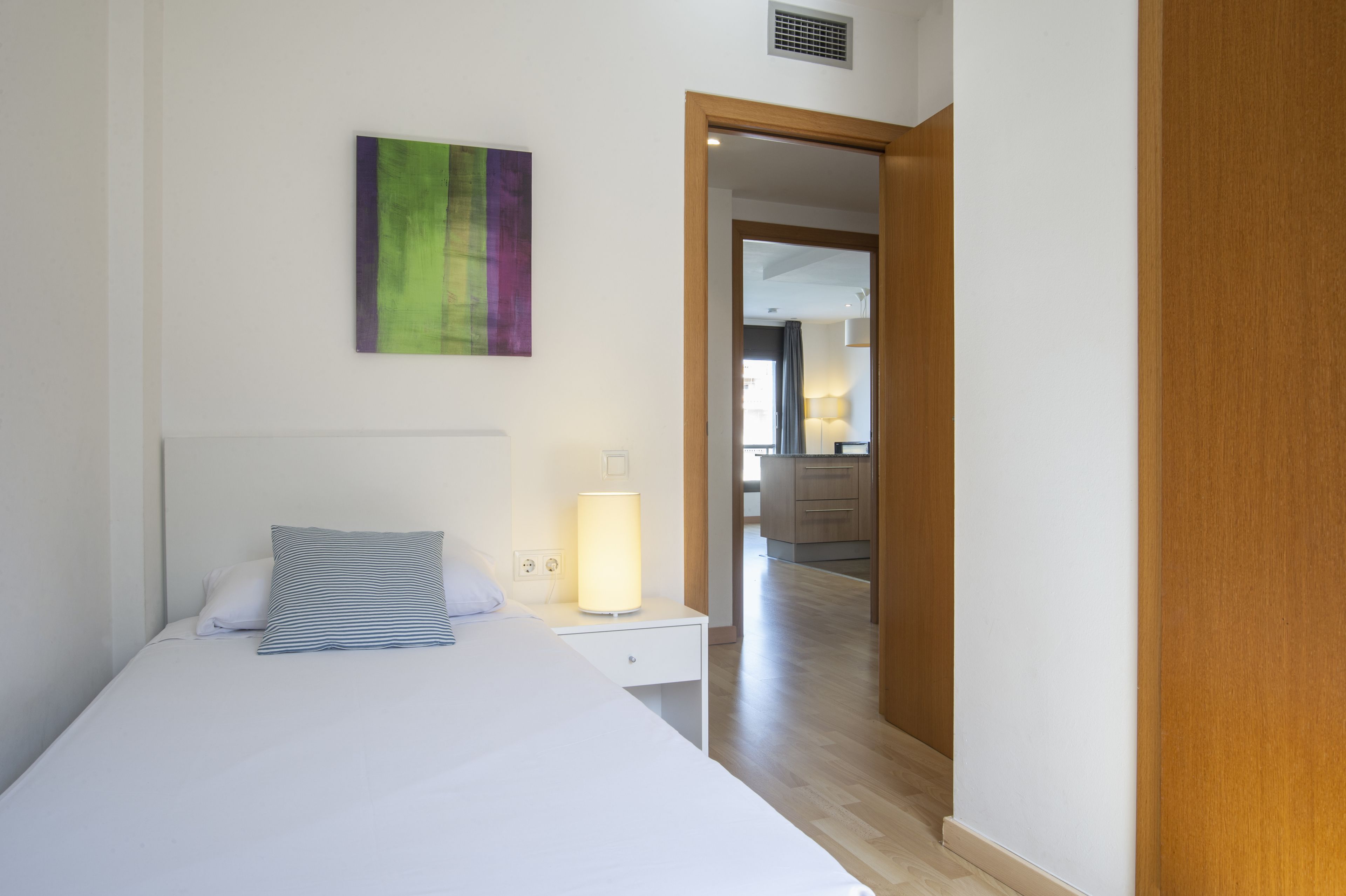 A bedroom with a bed, a bedside table with a lamp, a wall-mounted artwork, and a view into the adjacent kitchen area.