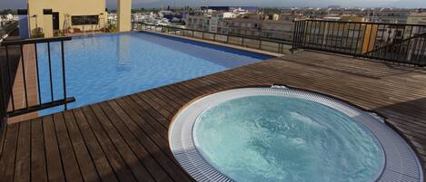 A rooftop pool with a hot tub, wooden deck, and a view of the cityscape.