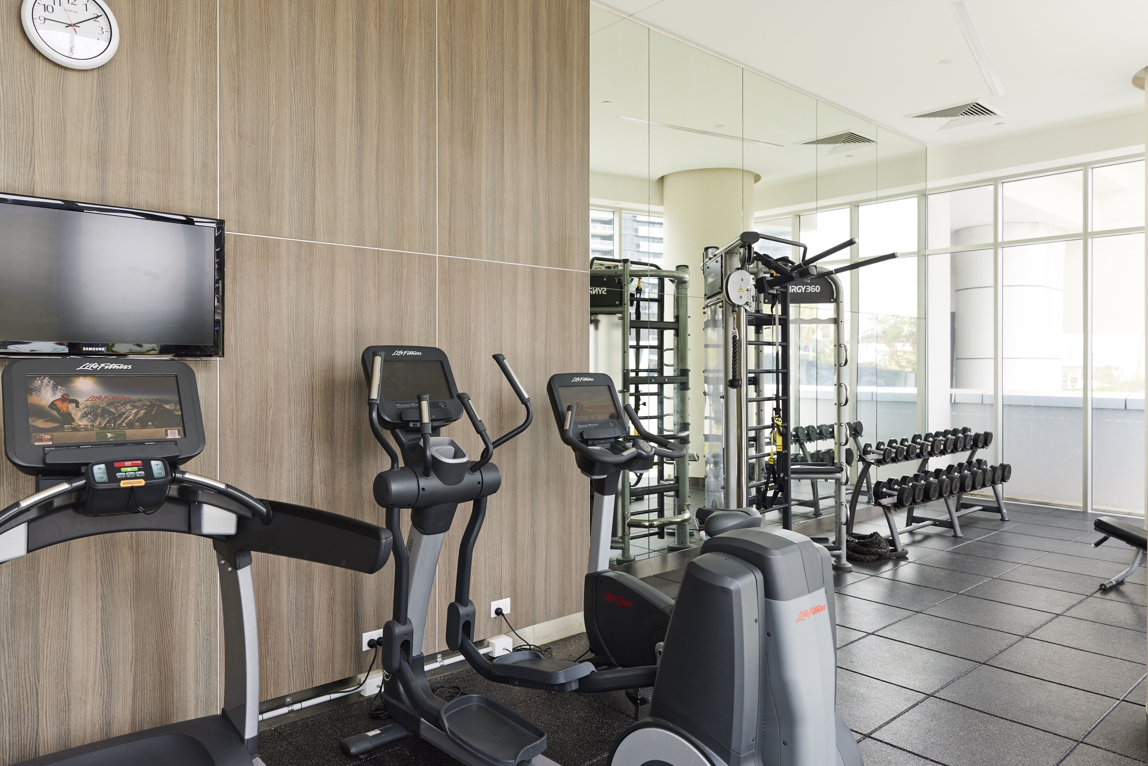A gym with cardio machines, weightlifting equipment, and a television screen displaying a workout video.